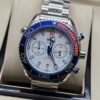 Omega — cronógrafo con esfera blanca, bisel azul con detalle naranja y brazalete de acero